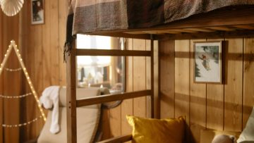 Przytulny pokój dziecięcy z drewnianymi ścianami i łóżkiem piętrowym, ozdobionym ciepłymi lampkami. Na łóżkach leżą koce, poduszki i pluszowe zabawki, a na dolnym łóżku otwarta książka. W tle fotel, dekoracje ścienne i nastrojowe oświetlenie tworzą ciepłą, domową atmosferę.