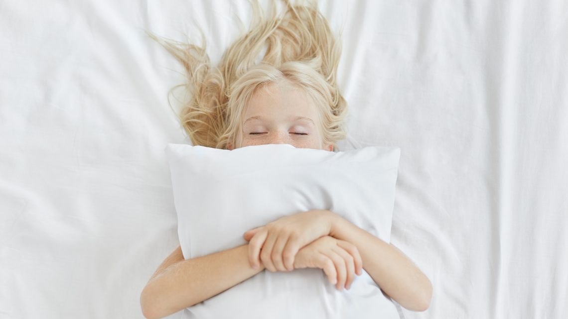 Beztroska, spokojna dziewczynka leżąca na białej pościeli, obejmująca poduszkę, mając jednocześnie przyjemne sny. Blondynka z piegami śpi w łóżku po spędzeniu całego dnia na pikniku. Spokojne dziecko