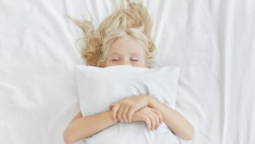 Beztroska, spokojna dziewczynka leżąca na białej pościeli, obejmująca poduszkę, mając jednocześnie przyjemne sny. Blondynka z piegami śpi w łóżku po spędzeniu całego dnia na pikniku. Spokojne dziecko